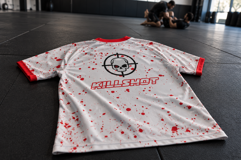 VHTK Killshot rash guard laid on a training mat, showing red splatter and skull artwork with two grapplers blurred in the background.