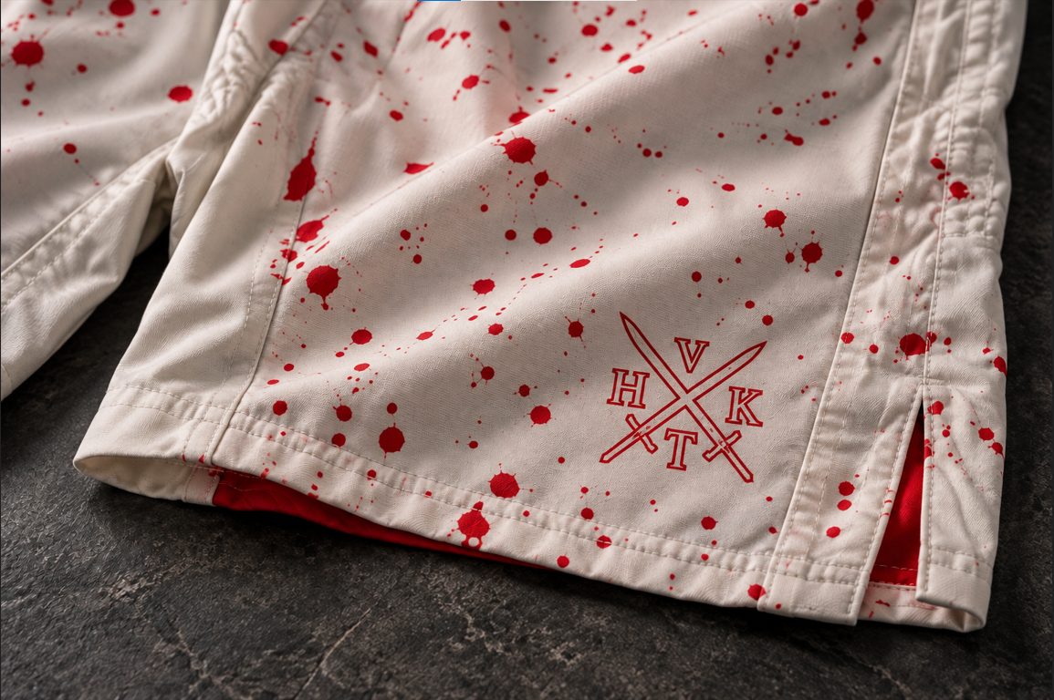Close-up of VHTK Killshot shorts showing red splatter artwork and VHTK branding on off-white fabric with a stitched leg hem.