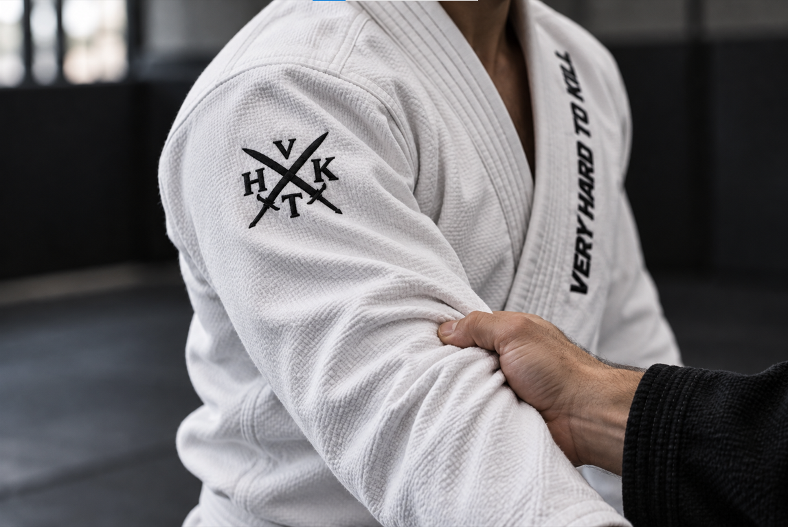 Partner gripping a white VHTK BJJ gi sleeve, showing reinforced shoulder stitching, woven fabric and the black embroidered emblem.