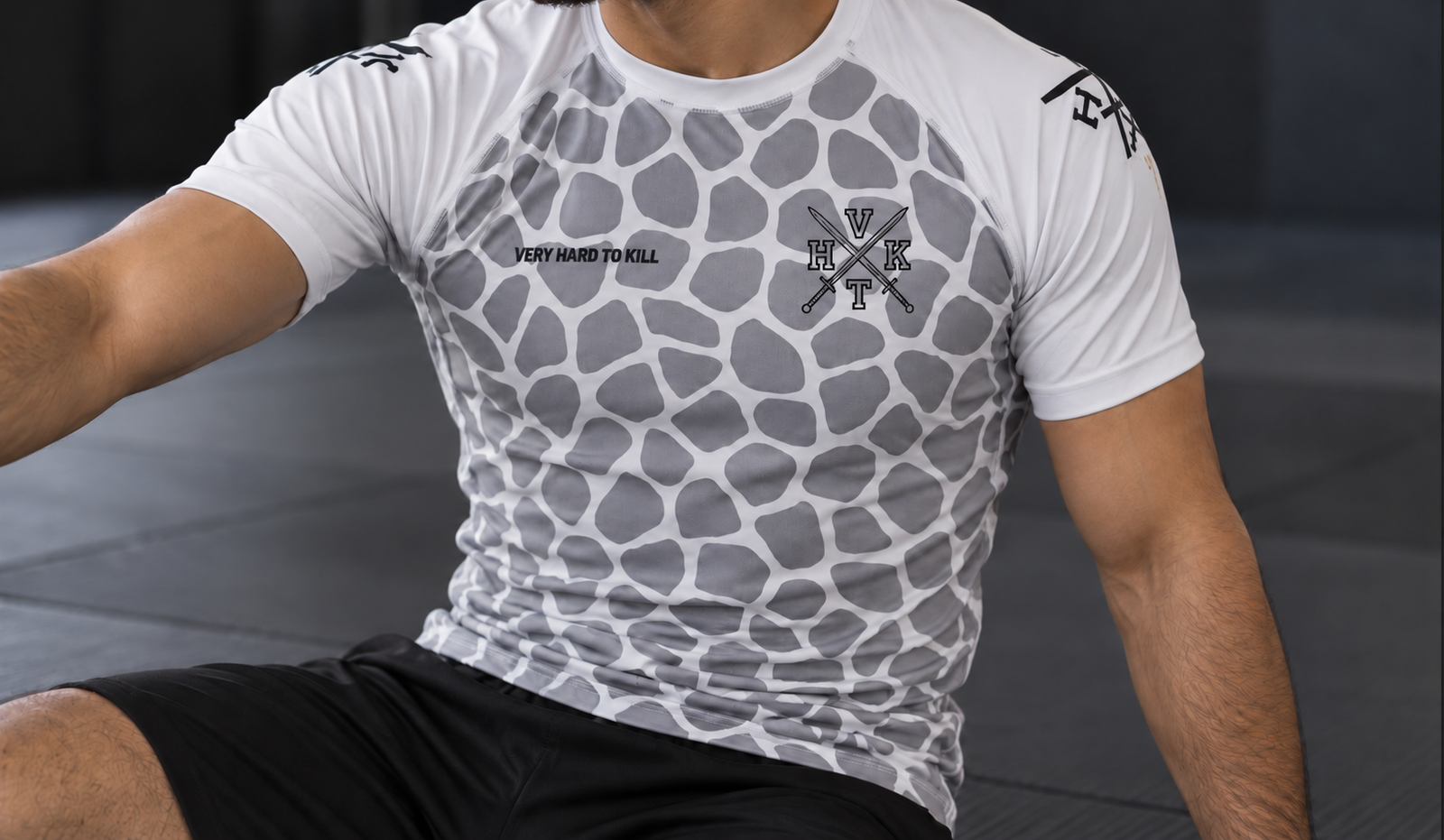 Athlete wearing a VHTK Giraffe 2.0 White BJJ rash guard with grey giraffe patterns and a crossed-swords chest emblem, seated on a grappling mat.
